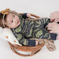 Smiling baby lying in a wicker basket wearing a green and brown camo romper with navy trim, as an adult zips open the inseam for an easy diaper change. Baby wears a mustard-colored bow headband.