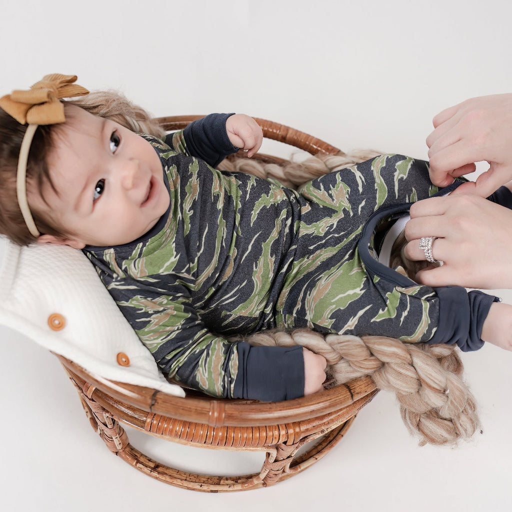 Smiling baby lying in a wicker basket wearing a green and brown camo romper with navy trim, as an adult zips open the inseam for an easy diaper change. Baby wears a mustard-colored bow headband.