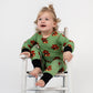 Toddler sitting in a vintage white rocking chair, wearing a green romper with black cuffs and a repeating tactical teddy bear print from 3rd Coast Limited.