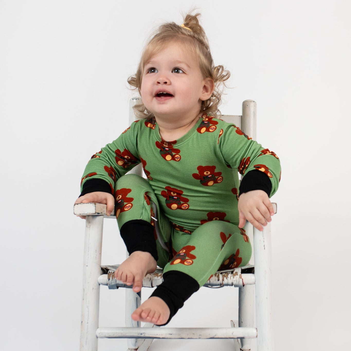 Toddler sitting in a vintage white rocking chair, wearing a green romper with black cuffs and a repeating tactical teddy bear print from 3rd Coast Limited.