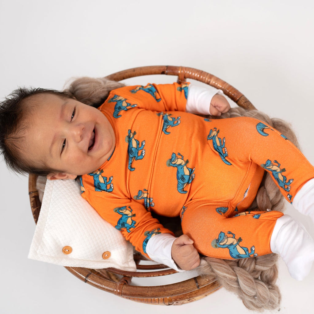 Smiling baby lying in a round wicker basket, wearing an orange romper with a blue T. rex print and white cuffs from 3rd Coast Limited.