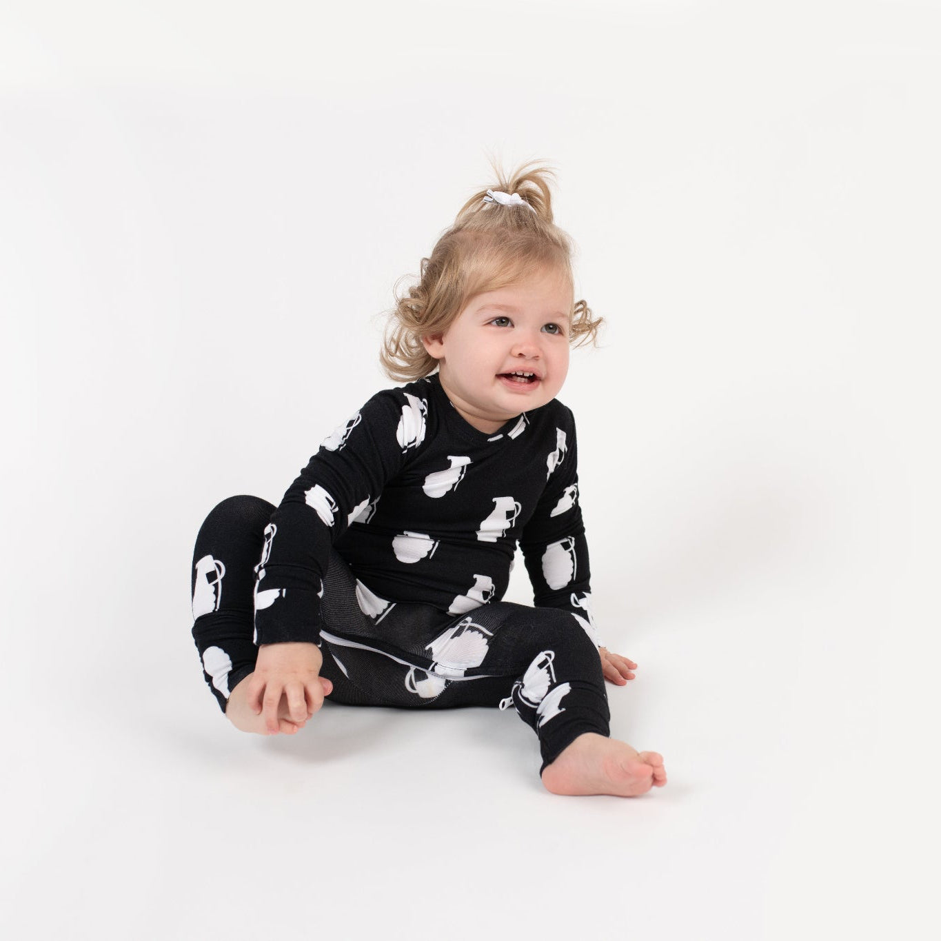 Smiling toddler sitting on a white backdrop, wearing a black long-sleeve romper with a white grenade pattern from 3rd Coast Limited.