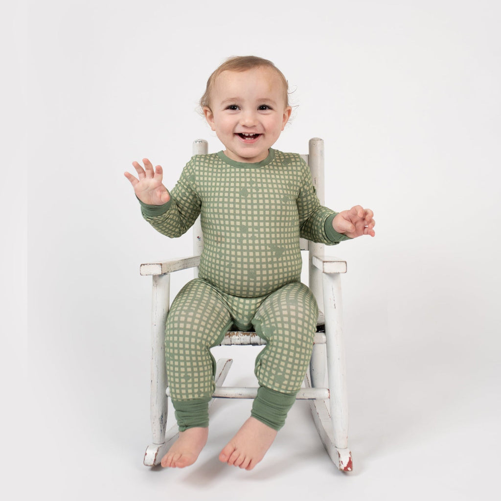 Smiling toddler sitting in a vintage white rocking chair, wearing a sage green romper with a tan grid pattern and matching green cuffs from 3rd Coast Limited.