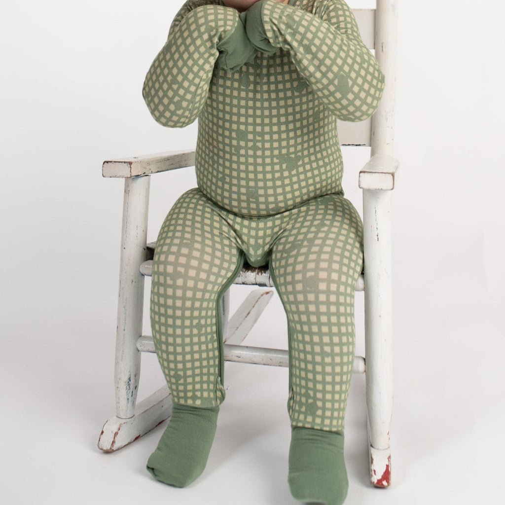 Smiling toddler sitting in a vintage white rocking chair, wearing a sage green romper with tan grid pattern, fold-over hand and foot cuffs covering hands and feet, from 3rd Coast Limited.
