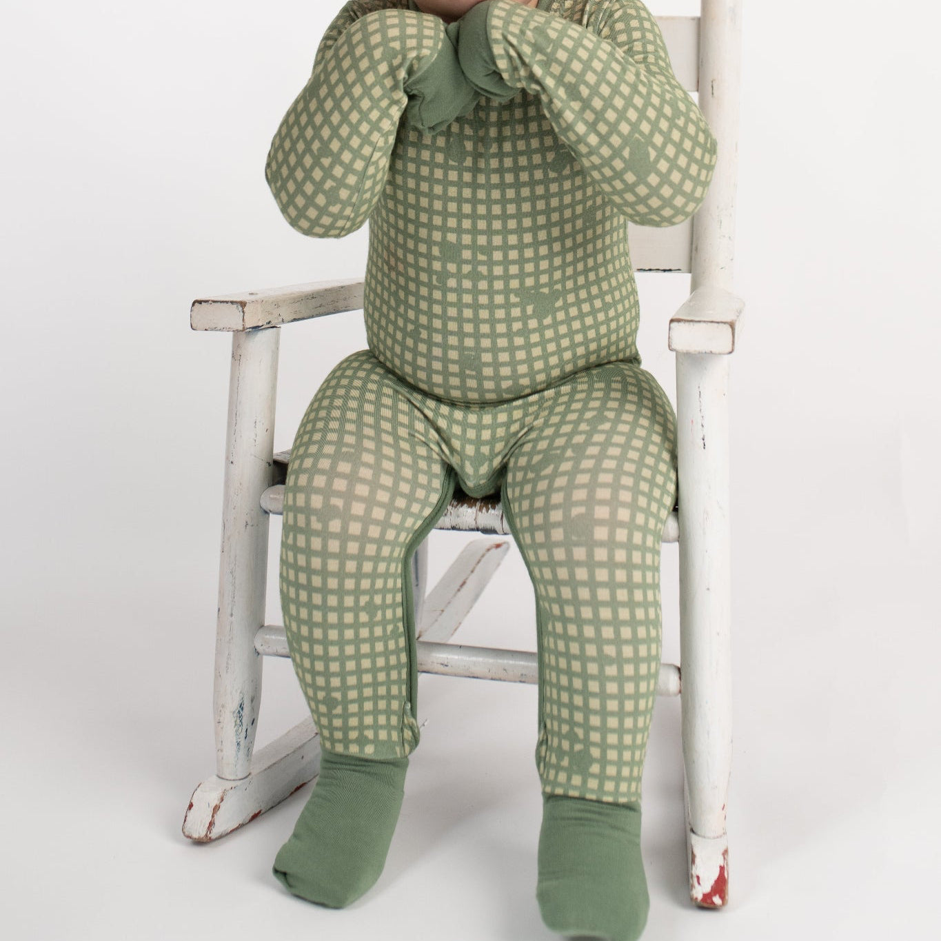 Smiling toddler sitting in a vintage white rocking chair, wearing a sage green romper with tan grid pattern, fold-over hand and foot cuffs covering hands and feet, from 3rd Coast Limited.