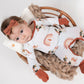 Baby lying in a round wicker basket on a chunky knit blanket, wearing a cream romper with rust cuffs, rainbow designs, and tactical unicorn print, paired with a rust-colored bow headband.