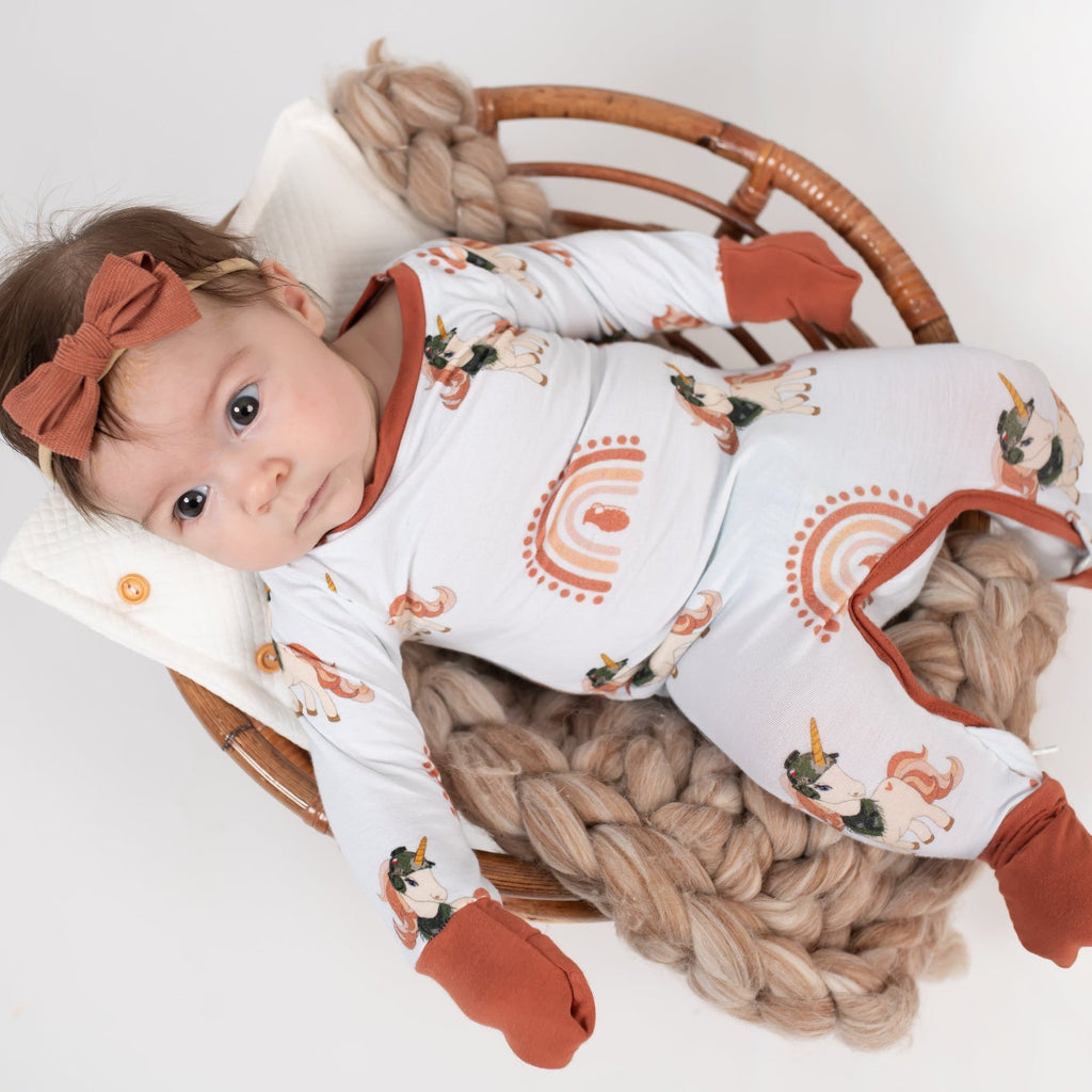 Baby lying in a round wicker basket on a chunky knit blanket, wearing a cream romper with rust cuffs, rainbow designs, and tactical unicorn print, paired with a rust-colored bow headband.