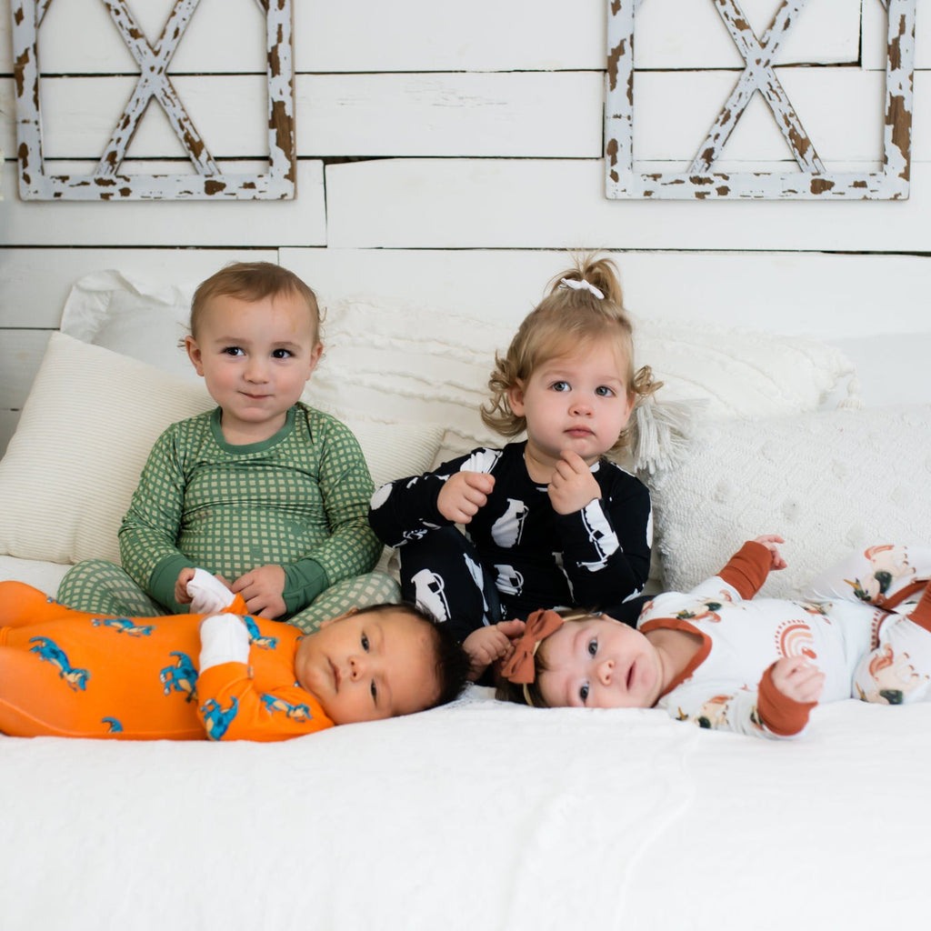 Four young children on a white bed in a farmhouse-style room, each wearing different printed pajamas—including green checkered, black with white grenades, bright orange with blue dinosaurs, and white with rust-colored rainbows and animals.