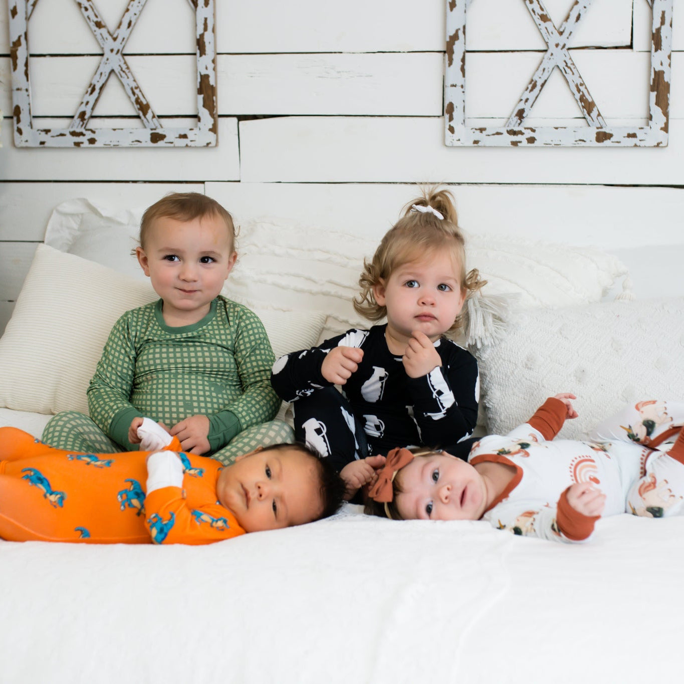 Four young children on a white bed in a farmhouse-style room, each wearing different printed pajamas—including green checkered, black with white grenades, bright orange with blue dinosaurs, and white with rust-colored rainbows and animals.