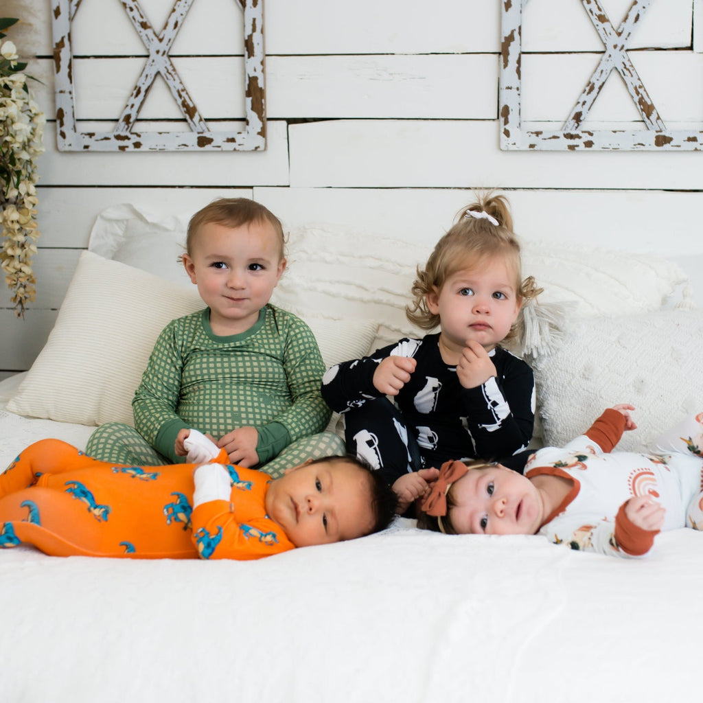 Four young children on a white bed in a farmhouse-style room, each wearing different printed pajamas—including green checkered, black with white grenades, bright orange with blue dinosaurs, and white with rust-colored rainbows and animals