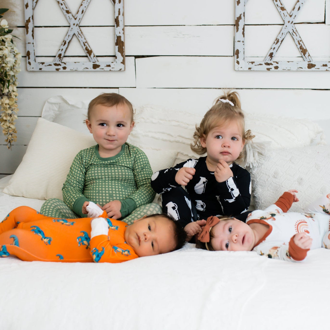 Four young children on a white bed in a farmhouse-style room, each wearing different printed pajamas—including green checkered, black with white grenades, bright orange with blue dinosaurs, and white with rust-colored rainbows and animals