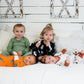 Four young children on a white bed in a farmhouse-style room, each wearing different printed pajamas—including green checkered, black with white grenades, bright orange with blue dinosaurs, and white with rust-colored rainbows and animals.