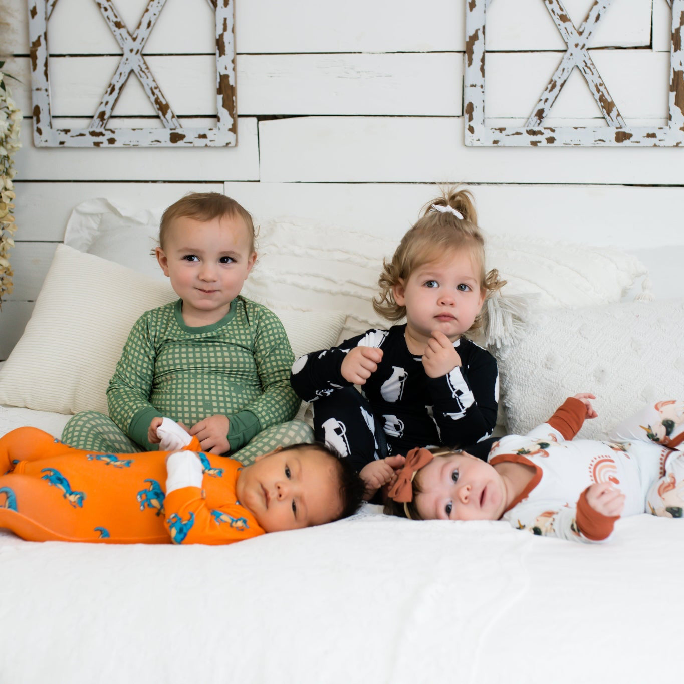 Four young children on a white bed in a farmhouse-style room, each wearing different printed pajamas—including green checkered, black with white grenades, bright orange with blue dinosaurs, and white with rust-colored rainbows and animals.