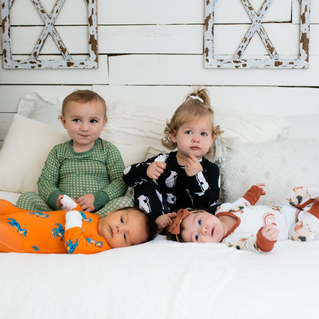 Four young children on a white bed in a farmhouse-style room, each wearing different printed pajamas—including green checkered, black with white grenades, bright orange with blue dinosaurs, and white with rust-colored rainbows and animals.