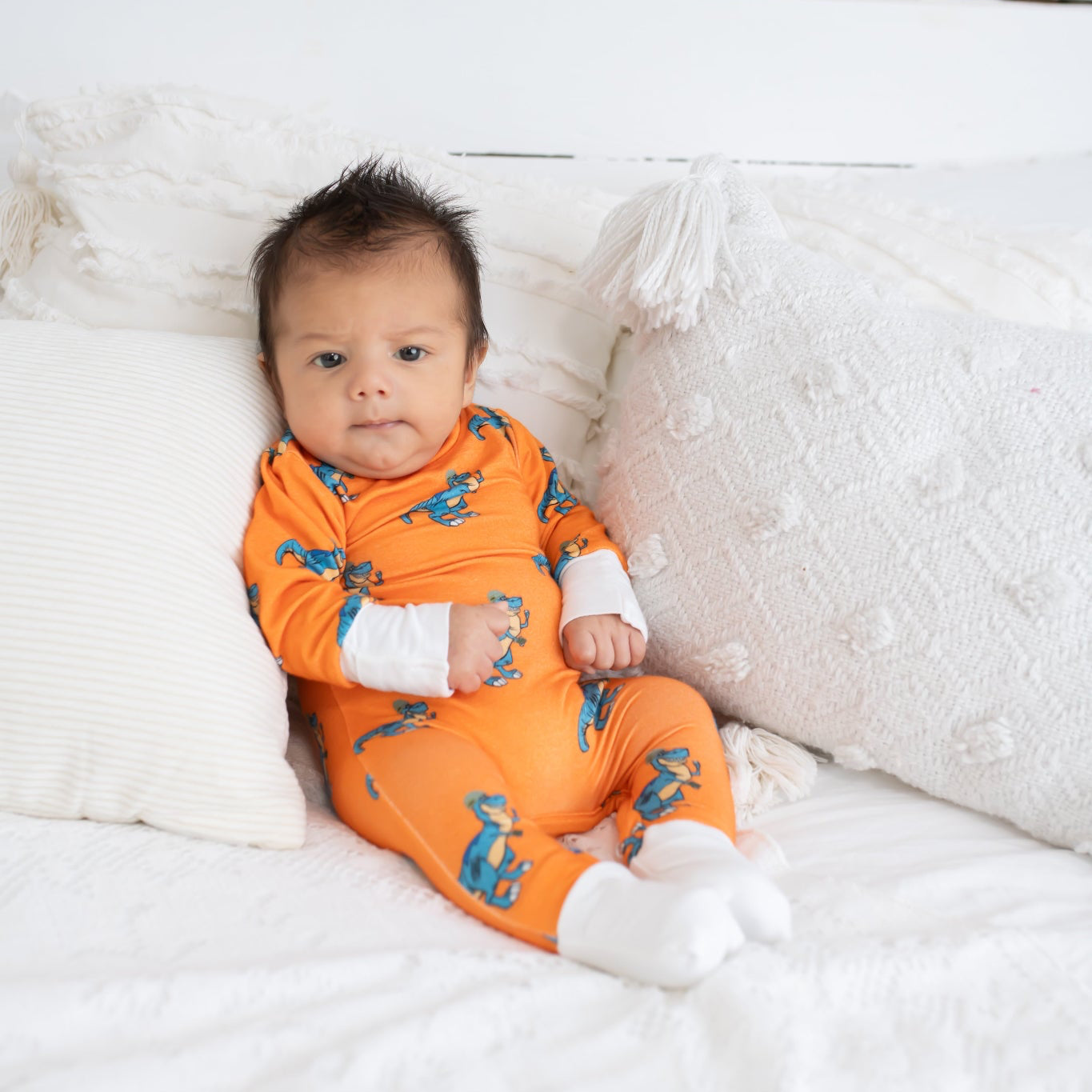 Baby sitting on a white bed with textured pillows, wearing an orange romper featuring a blue cartoon T. rex print and white cuffs from 3rd Coast Limited.