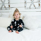 Toddler sitting on a white bed in a black romper with white grenade print, surrounded by textured white pillows and rustic farmhouse decor.