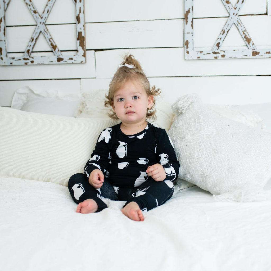 Toddler sitting on a white bed in a black romper with white grenade print, surrounded by textured white pillows and rustic farmhouse decor.