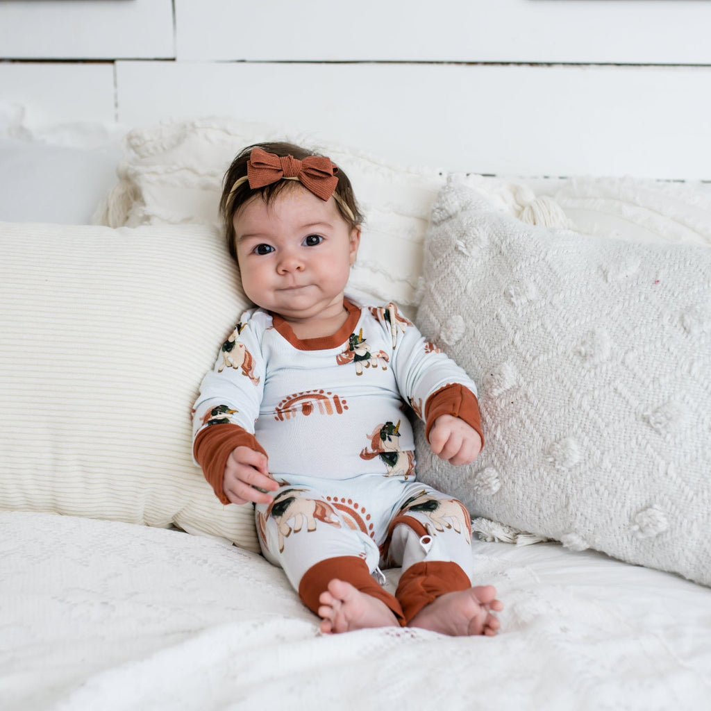 Baby sitting on a white bed with textured pillows, wearing a rust and cream-colored romper with rainbow and tactical print accents, and a matching rust bow headband.