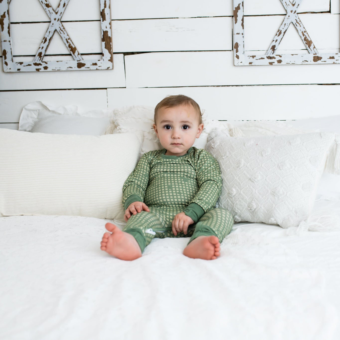 Toddler sitting on a white bed with textured pillows and farmhouse decor, wearing a sage green romper with a light tan grid pattern and matching green cuffs from 3rd Coast Limited.