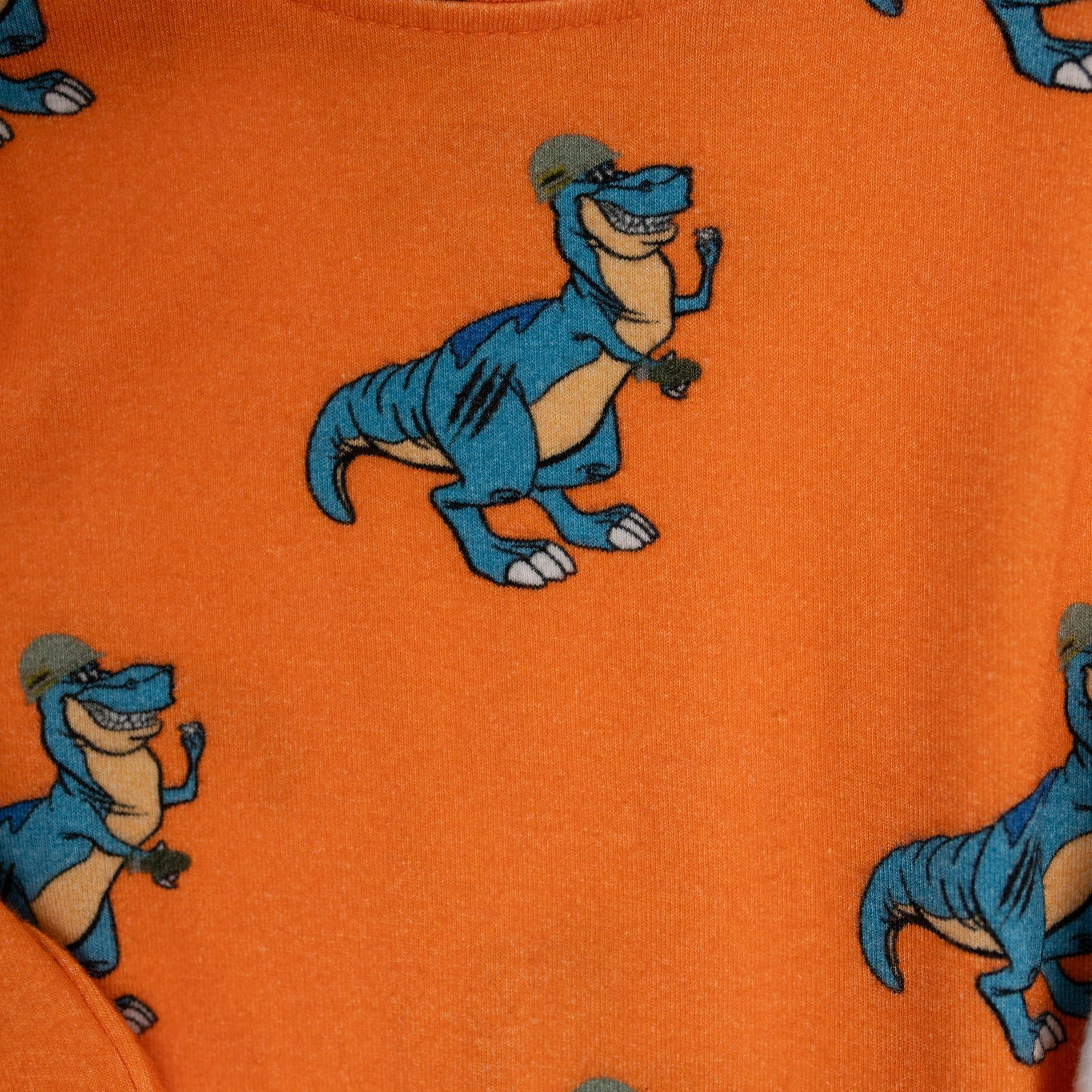 Close-up of an orange baby romper from 3rd Coast Limited, featuring a repeating print of a smiling blue T. rex wearing a military helmet and dog tags, flexing its arm.